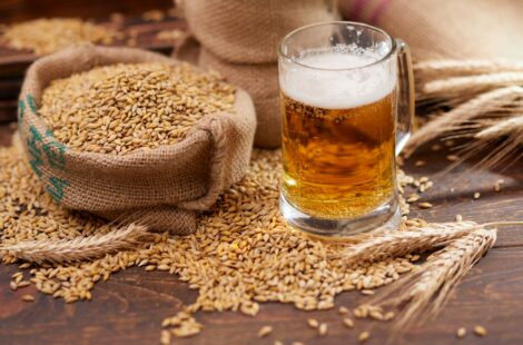 A glass of beer sitting on a wooden table next to an overflowing bag of grains.