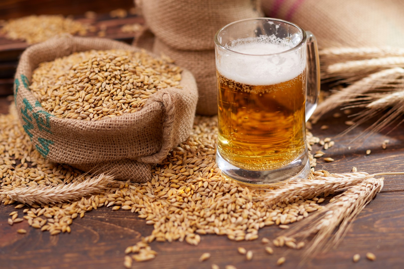 A glass of beer sitting on a wooden table next to an overflowing bag of grains.