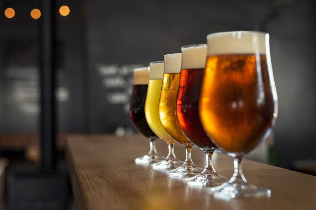 Five glasses on a wooden bar filled with different types of beer.